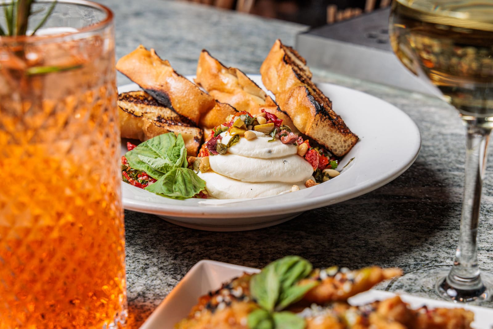 Burrata with grilled bread, basil and chopped tomato on a white plate, a spritz in a tall glass beside it