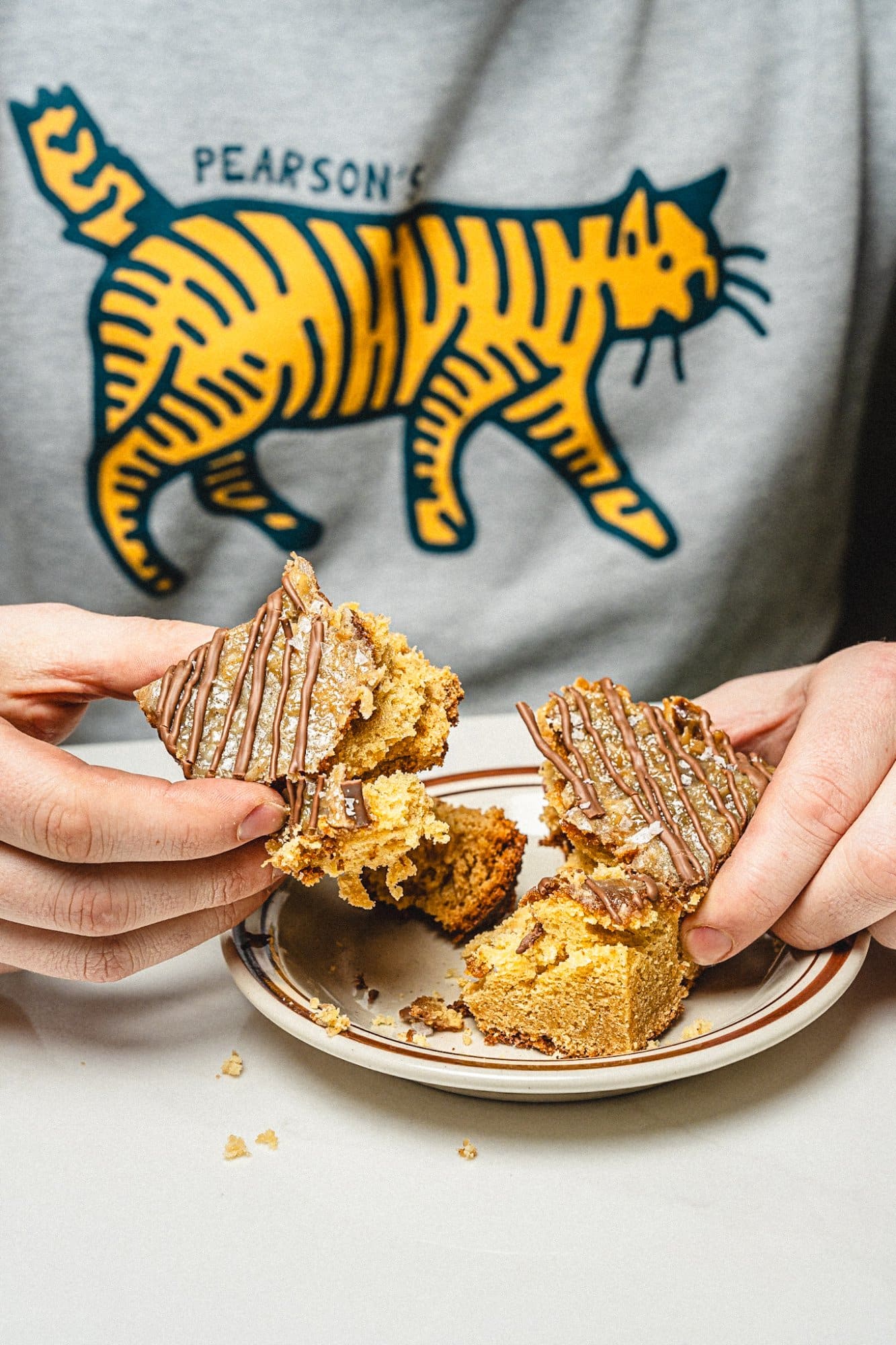 Hands breaking apart a chocolate-drizzled bar on a plate, a Pearson's cat sweatshirt behind